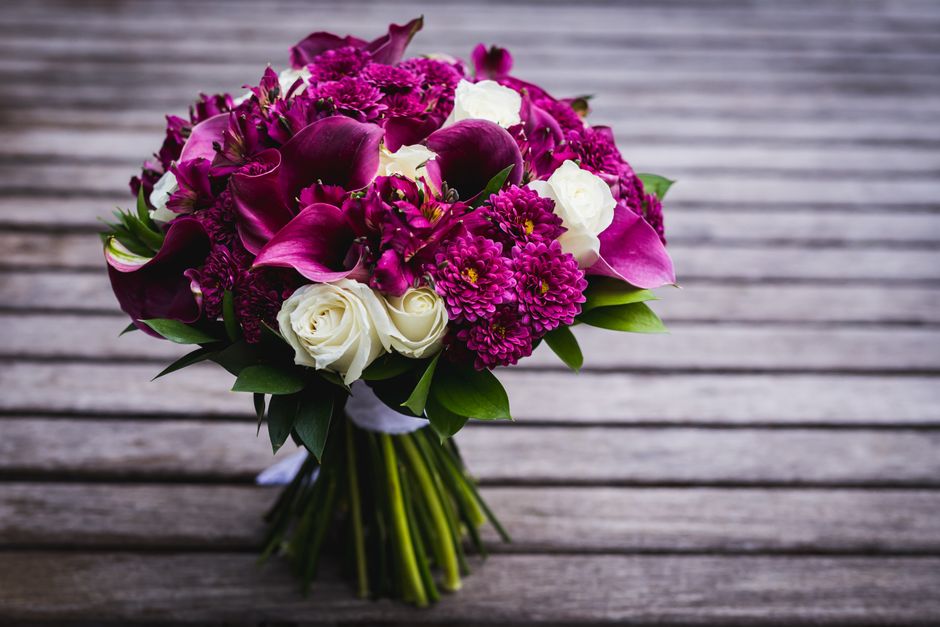 A vibrant bouquet of fresh flowers arranged with deep magenta calla lilies, bright pink chrysanthemums, purple alstroemeria, and elegant white roses, all nestled in lush green foliage. The bouquet is 