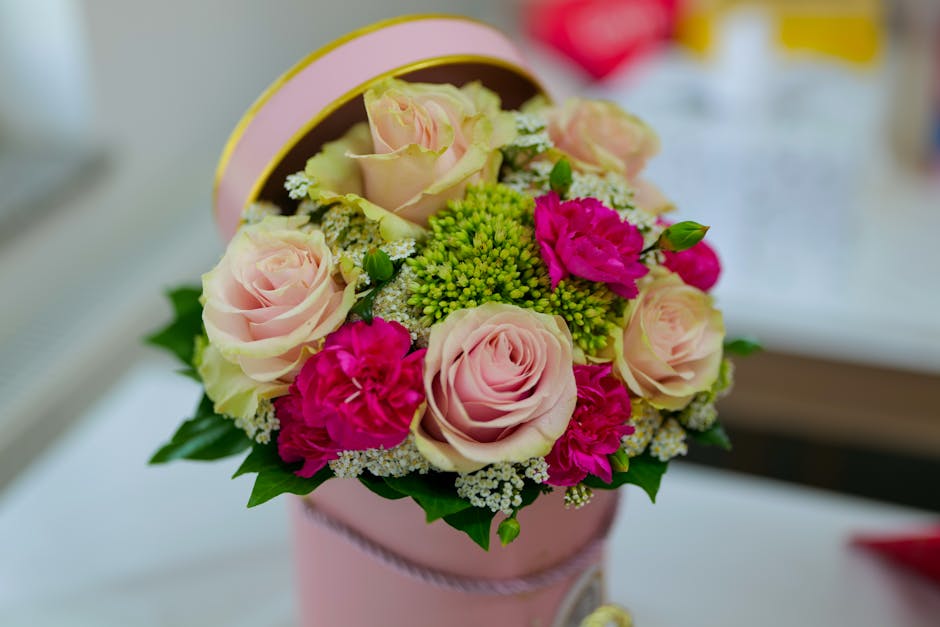A round floral arrangement inside a pink cylindrical box featuring pale pink roses, bright pink carnations, and clusters of green hypericum berries, accented with tiny white filler flowers and lush gr