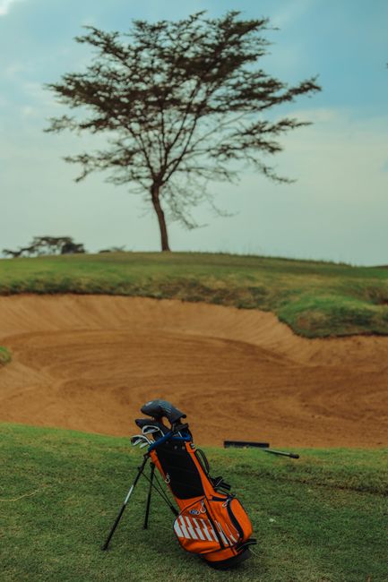 A vibrant orange and black golf bag with multiple compartments and a shoulder strap is positioned on the edge of a well-manicured green golf course near a sand bunker. The bag contains several golf cl