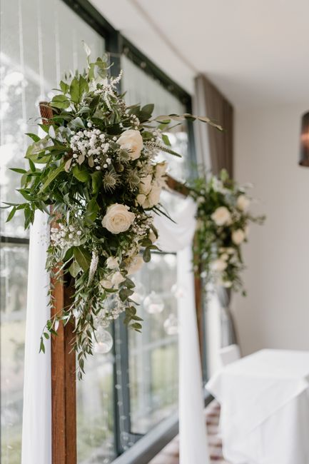 A floral arrangement featuring a lush cascade of white roses, baby's breath, and various green foliage, draping over a wooden stand as part of an elegant décor for a wedding or special event. The bou