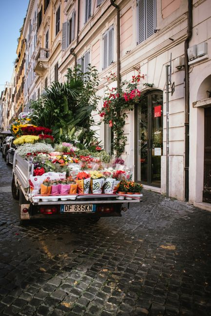 A delivery truck parked on a cobblestone street outside a building with white walls and tall windows, overflowing with a variety of fresh flowers arranged in bouquets and large floral displays. The fl