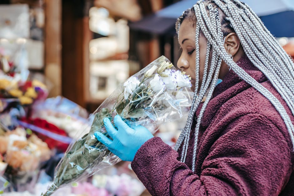 A woman with silver braided hair wearing a maroon fleece jacket and blue gloves is holding a bouquet of white flowers wrapped in clear plastic. She is inspecting or smelling the flowers in an outdoor 