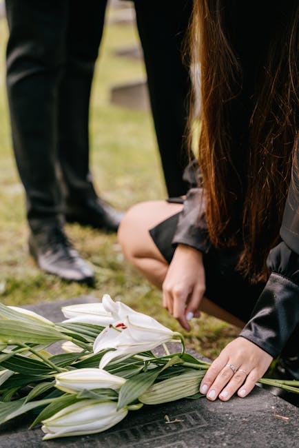 A person kneeling at a grave site, arranging a floral bouquet consisting of white lilies with large, elegant petals and prominent stamens, surrounded by broad green leaves. The arrangement is placed o
