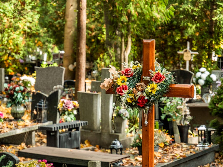 A wooden cross decorated with a floral arrangement featuring red roses, yellow daisies, pale pink flowers, and green foliage, situated amidst a graveyard with various headstones and memorial plaques. 