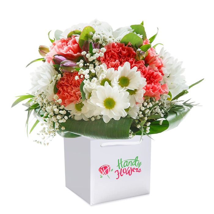 Perfectly poised against a seamless white backdrop, a lush floral display rises from a structured, square white gift bag, its form defined by both elegance and joyful abundance. At first glance, the eye is drawn to the cacophony of colour and texture: coral and peach carnations unfurl their ruffled petals in soft, rippling waves, while vibrant red accents inject a spirited energy reminiscent of Chigwell's lively community events. Snowy white daisies, their golden-green centers glowing brightly, mirror the light-hearted cheer of open spaces along Hainault Road. Wisps of white gypsophila (baby's breath) punctuate the scene with delicate grace, their tiny blossoms forming a lace-like veil between bolder blooms. Subtle unopened buds tinged with purple suggest the quiet promise of gardens waking in the early hours, perhaps inspired by morning walks along Chigwell's tranquil lanes. Wraps of deep green, broad-leaved foliage envelop the bouquet's base, creating a natural, glossy frame that highlights each flower's individual beauty and the harmonious composition as a whole. The entire arrangement is composed with intuitive care and expertise-a full, rounded silhouette that speaks of seasoned hands and an eye for local charm. Ideal for celebrating a special occasion or marking life's everyday joys, this bouquet offers a ready-to-gift sense of occasion, resonate with Chigwell's welcoming spirit. It's a testament to the care found in local flower shops, inviting recipients to enjoy freshness and artistry delivered right to their door.
