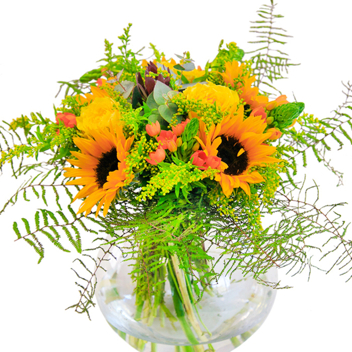 As the eye lingers over this sumptuous bouquet, the vibrant essence of Chigwell's open spaces is brought to life in a symphony of sun-kissed colour. Dominating the arrangement are broad, golden-yellow sunflowers, their textured petals fanning out in radiant circles, each center dark and velvety, hinting at late summer days in the countryside bordering Chigwell. Interlaced with these joyful blooms, the tightly wound petals of yellow garden roses invite closer admiration, each one expertly opened to reveal softness and intricate spirals reminiscent of local cottage gardens along Manor Road. Glossy red Hypericum berries glisten among the foliage, adding tangible bursts of energy and playfully contrasting against the dominant yellows. The foliage is lush and varied: delicate fronds of fern cascade gently, introducing airy lines and natural movement, while slender spikes of light green filler such as goldenrod nestle in, dotted with miniature yellow blossoms. Subtle touches of bluish eucalyptus infuse the bouquet with a cool, refreshing hint, and every stem stands cradled in a crystal-clear, spherical vase. The water within reflects the afternoon light, enhancing the bouquet's vivid hues and the freshness for which Chigwell's flower shops are known. This creation is a heartfelt choice for anniversaries, housewarmings, or simply celebrating the warmth of the community. Each element speaks to the skillful hand of Chigwell's florists, echoing the town's blend of natural beauty and vibrant spirit.