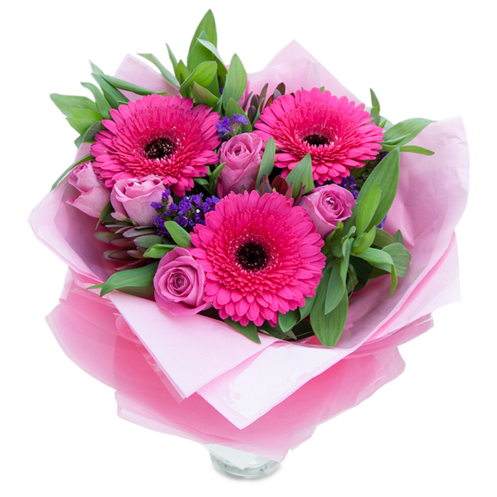 This bouquet reads like a friendly, pink-hued embrace: three striking fuchsia gerbera daisies command attention with their layered petals and deep, dark centres, while a ring of lavender-pink roses nestle close, petals tightly rolled in the way of newly cut, long-lasting blooms. Small sprigs of purple statice are scattered thoughtfully through the design, lending a textured, almost meadow-like counterpoint to the more formal roses. The greenery-full, glossy leaves that suggest salal and rounded eucalyptus-provides a cool, restorative contrast and holds the composition together with a natural fullness. The flowers are wrapped as part of their story: an inner pale-pink tissue paper creates a soft cushion, and over it a crinkled, translucent fuchsia-pink cellophane gives the bouquet a slight sheen and a celebratory finish; careful folding around the stems reveals the clear glass vase's rim so the recipient can set the arrangement immediately on display. In gentle daylight the colours glow with optimism, the roses offering a faint sweet perfume and the gerberas a subtle green freshness that lifts the senses. As a florist familiar with Chigwell and the surrounding towns, I imagine this arrangement brightening a kitchen table after a walk in Epping Forest or greeting a loved one on a special anniversary or birthday-an uncomplicated, expertly composed expression of warmth and appreciation.